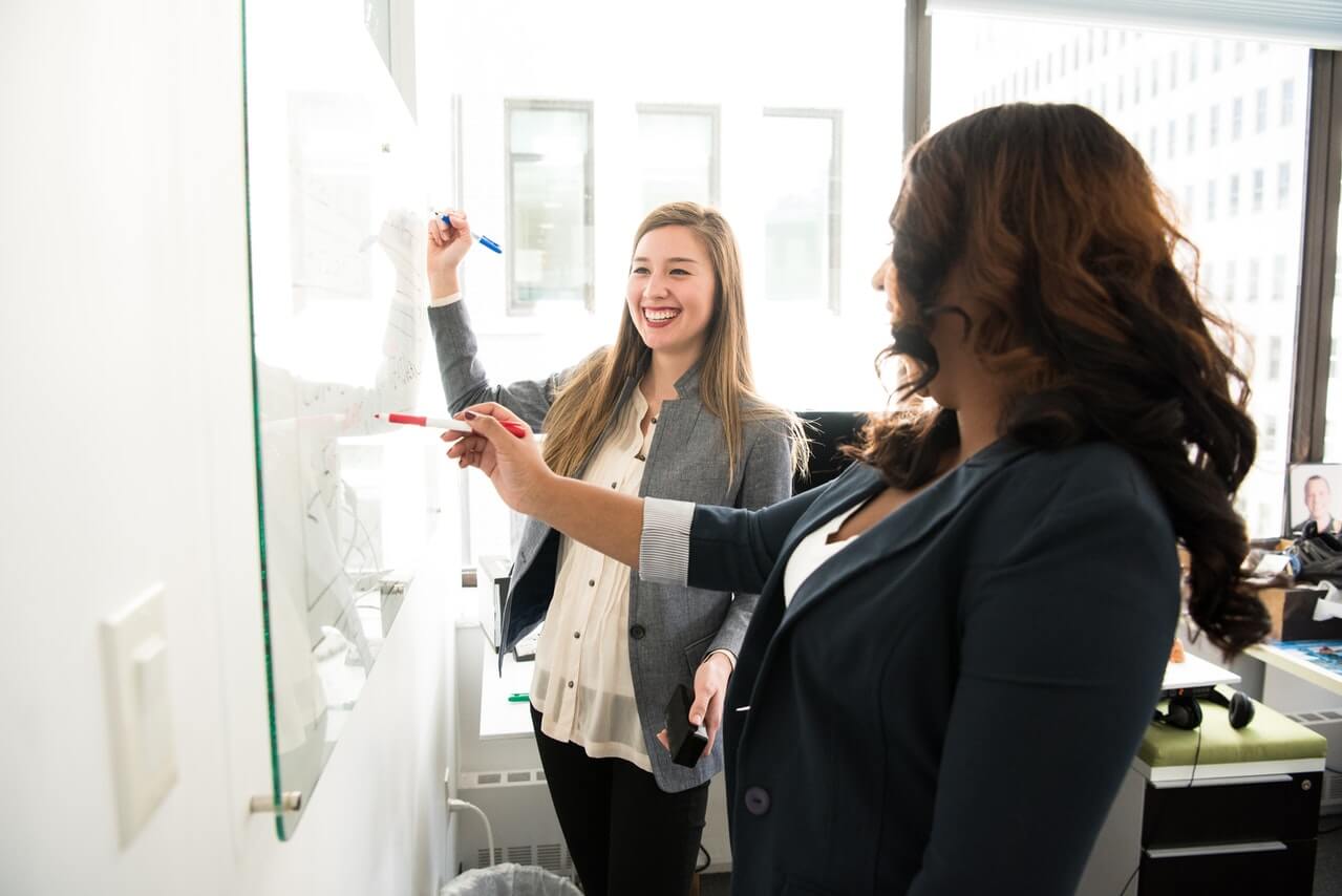Two women laughing in front of a whiteboard