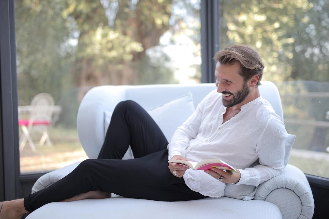 Man in White Long Sleeves and Black Pants Sitting on Sofa While Holding a Book