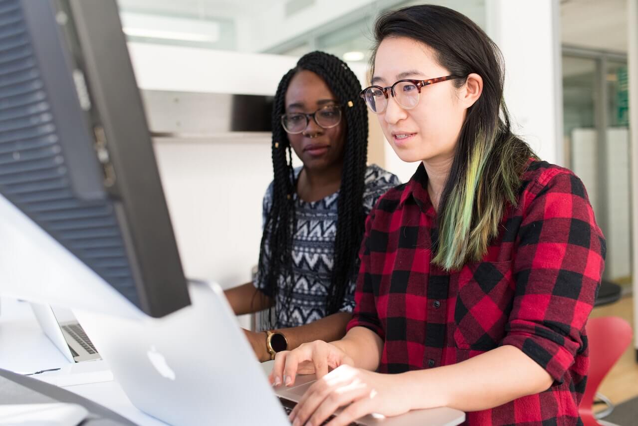 Woman in checkered shirt teaching colleague using a macbook