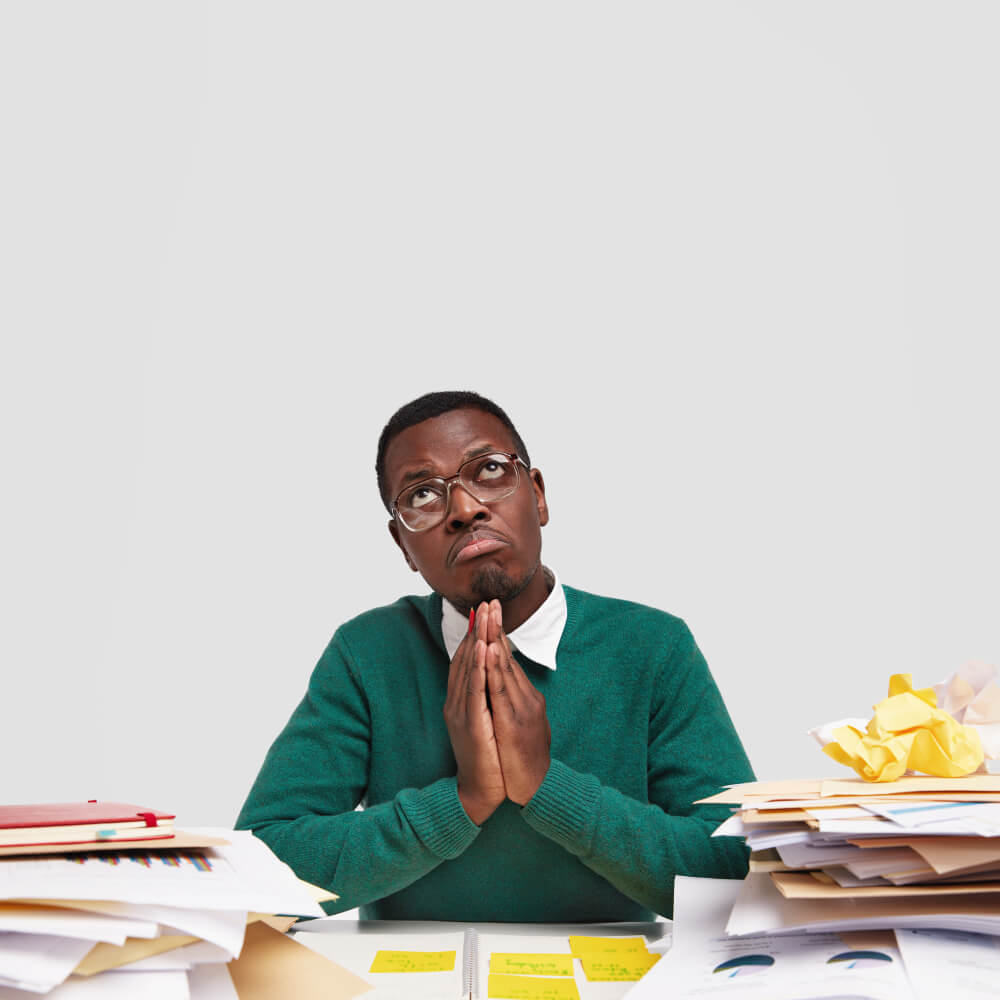 attractive-black-man-keeps-hands-praying-gesture-prays-while-works-desktop-wears-transparent-glasses-looks-with-pity-expression